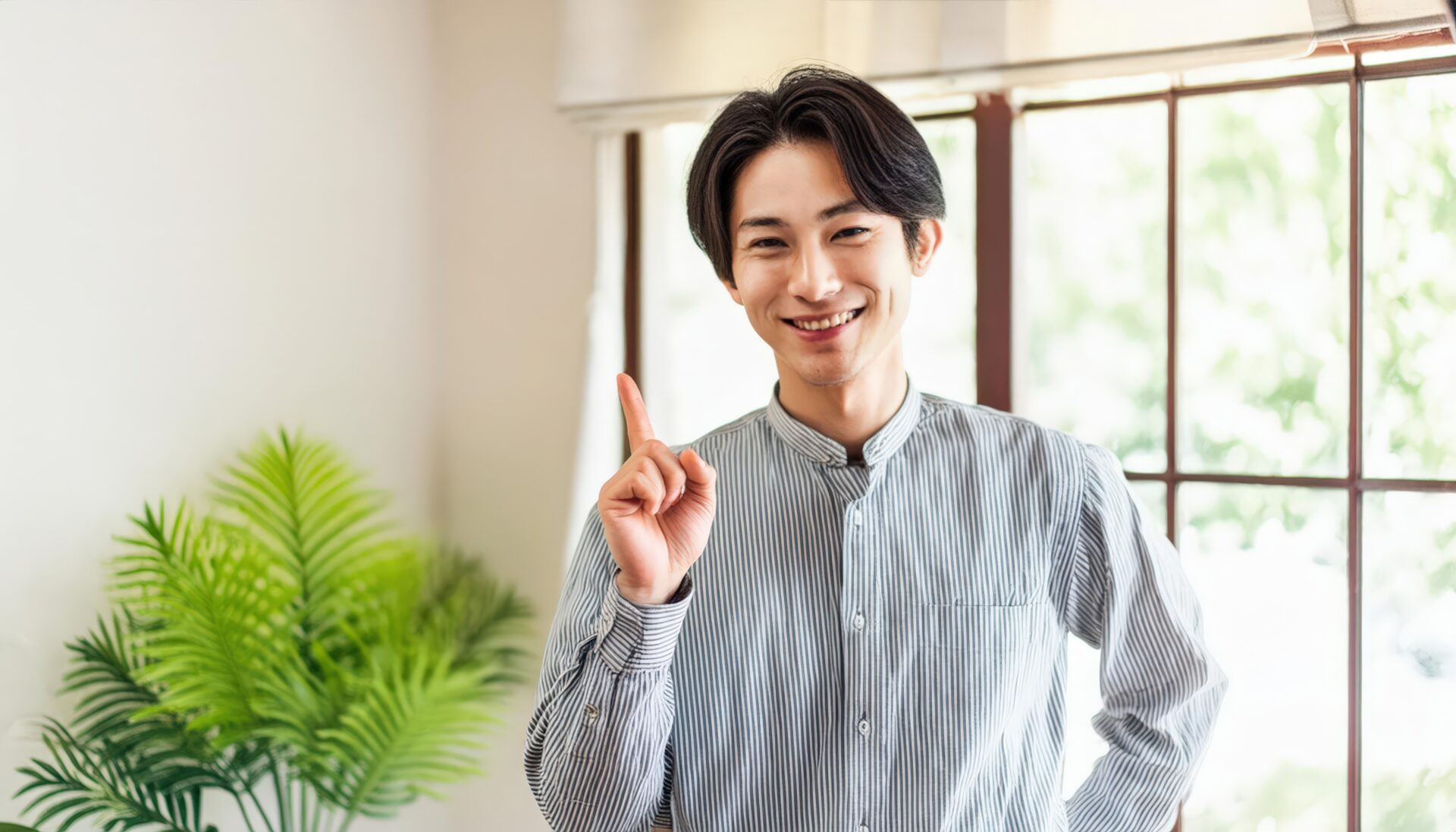 室内で人差し指を立てる笑顔の若い男性（窓とグリーン）