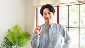 室内で人差し指を立てる笑顔の若い男性（窓とグリーン）