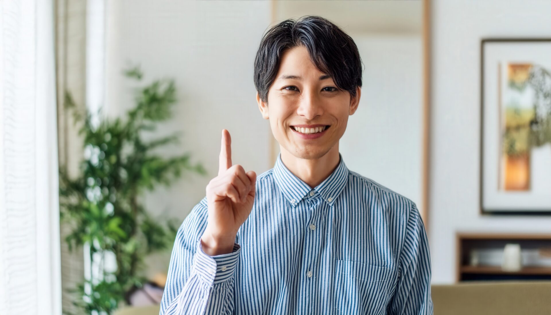 室内で人差し指を立てる笑顔の若い男性（ストライプシャツ・アートフレーム）