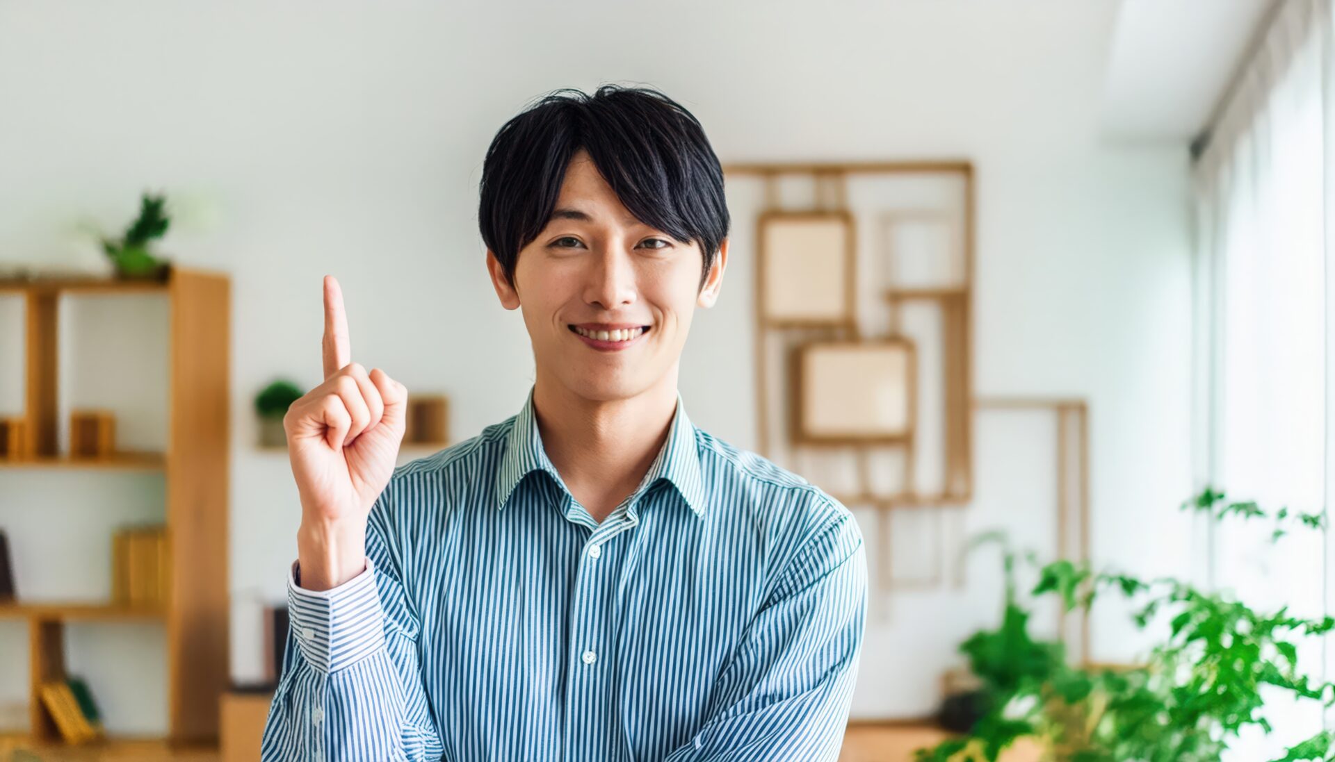 室内で人差し指を立てる笑顔の若い男性（ストライプシャツ・棚）
