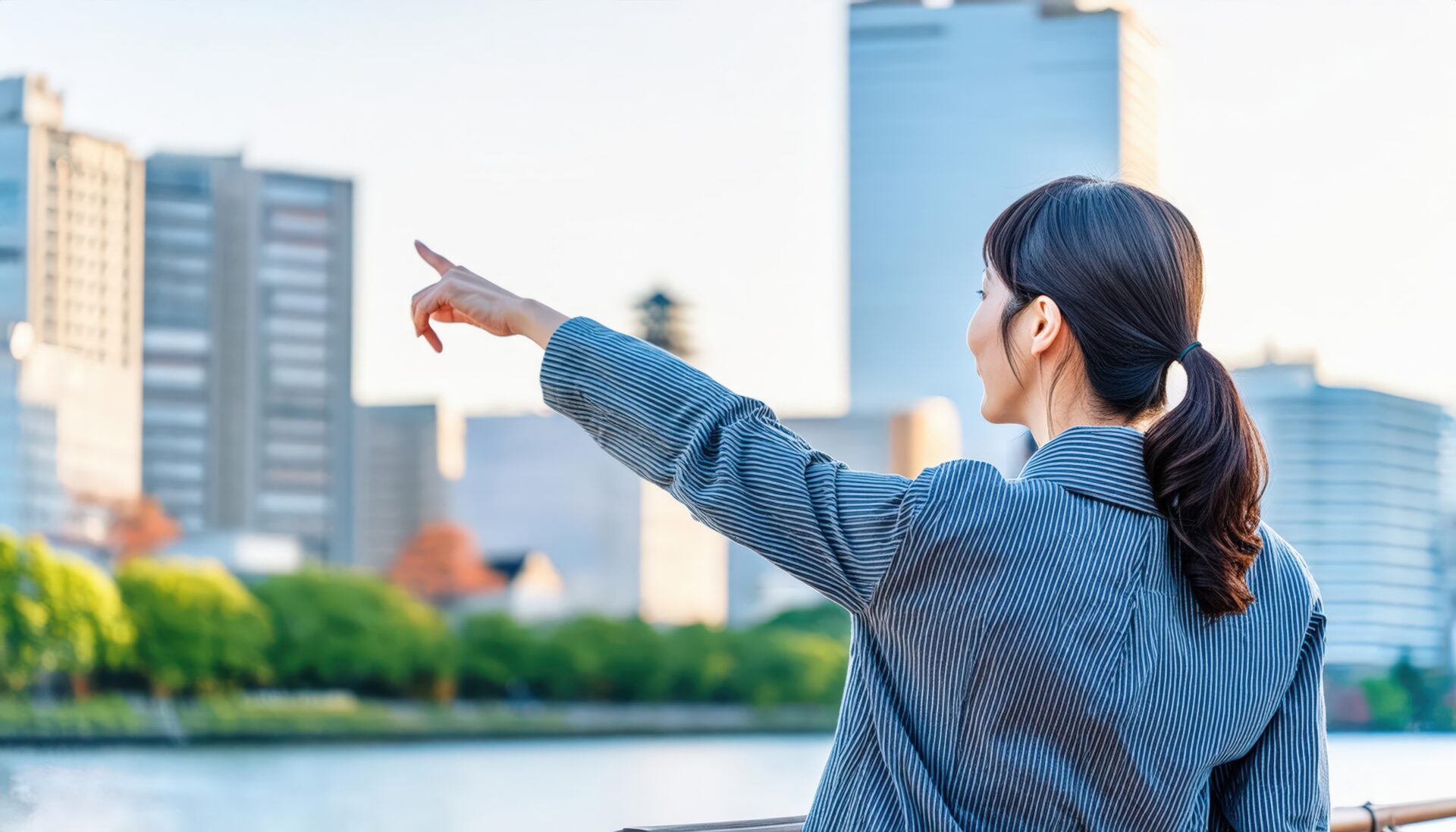 ストライプスーツを着た後ろ姿の若い人が川沿いで指をさす