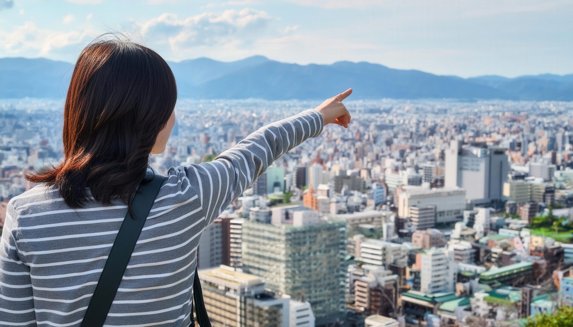 ストライプのトップスを着た後ろ姿の若い人が高台から街を指す