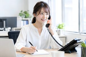 オフィスの固定電話に笑顔で応対しメモを取る20代女性