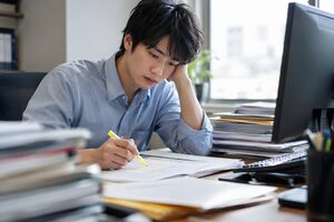 書類の山を前に蛍光ペンで確認作業をする20代男性