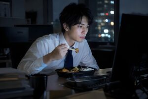 夜のデスクでコンビニ弁当を食べながら作業を続ける20代男性