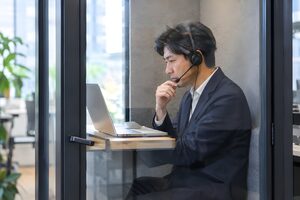 オフィスの個室ブースでヘッドセットを着けてオンライン会議をする40代男性