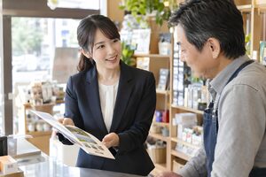 小さな店のカウンターで店主にパンフレットを説明する20代の営業女性