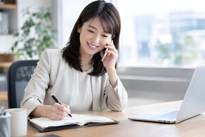 電話でアポイントを取り手帳に予定を書き込む30代の営業女性