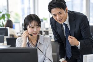 若手の電話営業を横で聞いて指導する40代の営業マネージャー