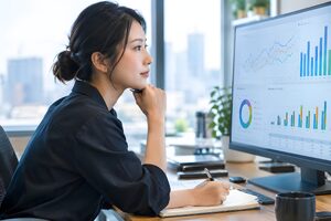 ダッシュボードを映した大型モニターを見ながらメモを取る30代の女性アナリスト