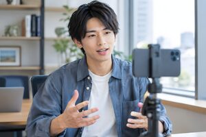 オフィスで三脚に立てたスマートフォンに向かって縦動画を撮る20代男性