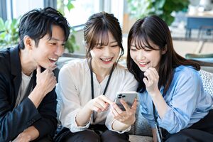 会社の投稿が表示されたスマートフォンを囲んで喜ぶ3人のマーケティングチーム