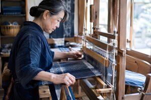 木製の手織り機で藍と白の布を織る60代の女性