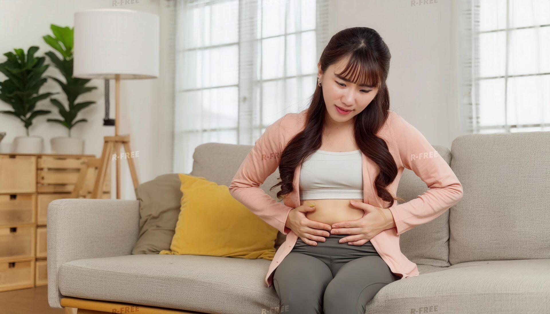 ソファでお腹を見つめ両手で押さえるピンクカーディガンの女性