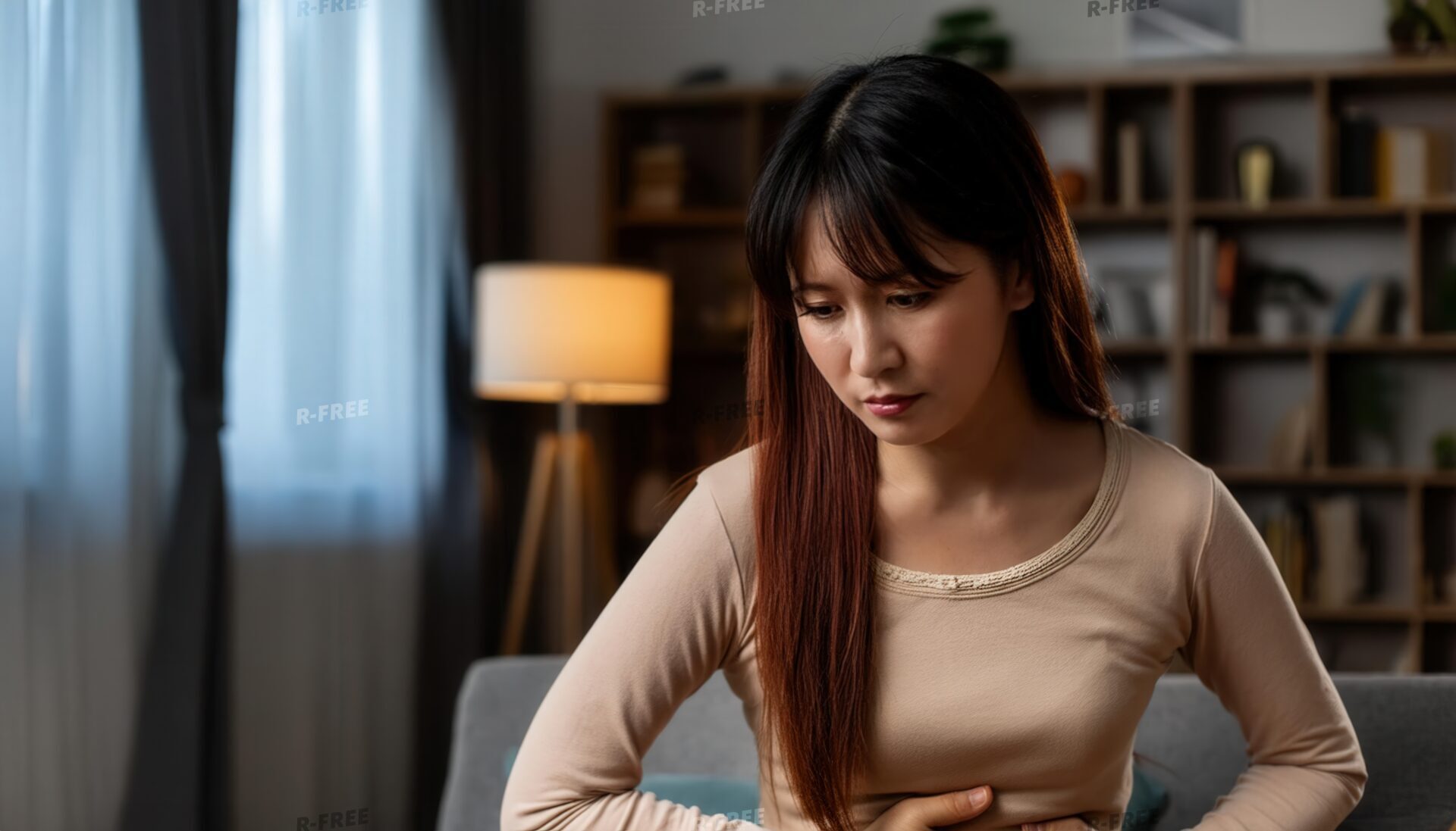 夜のソファでお腹を抱えて苦しそうにするベージュ長袖の女性