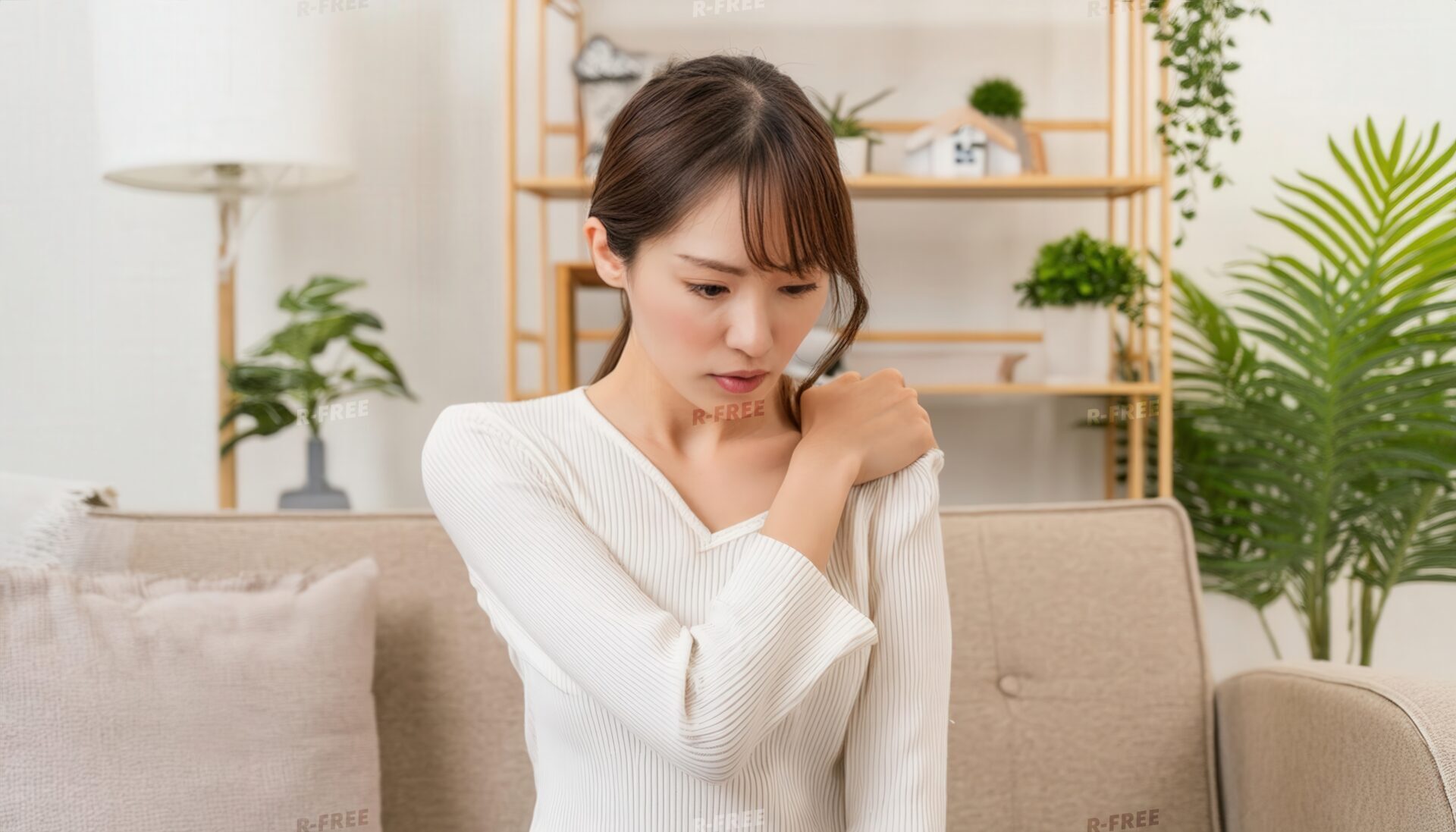 肩こりで左肩を押さえ下を向く白いVネックの女性