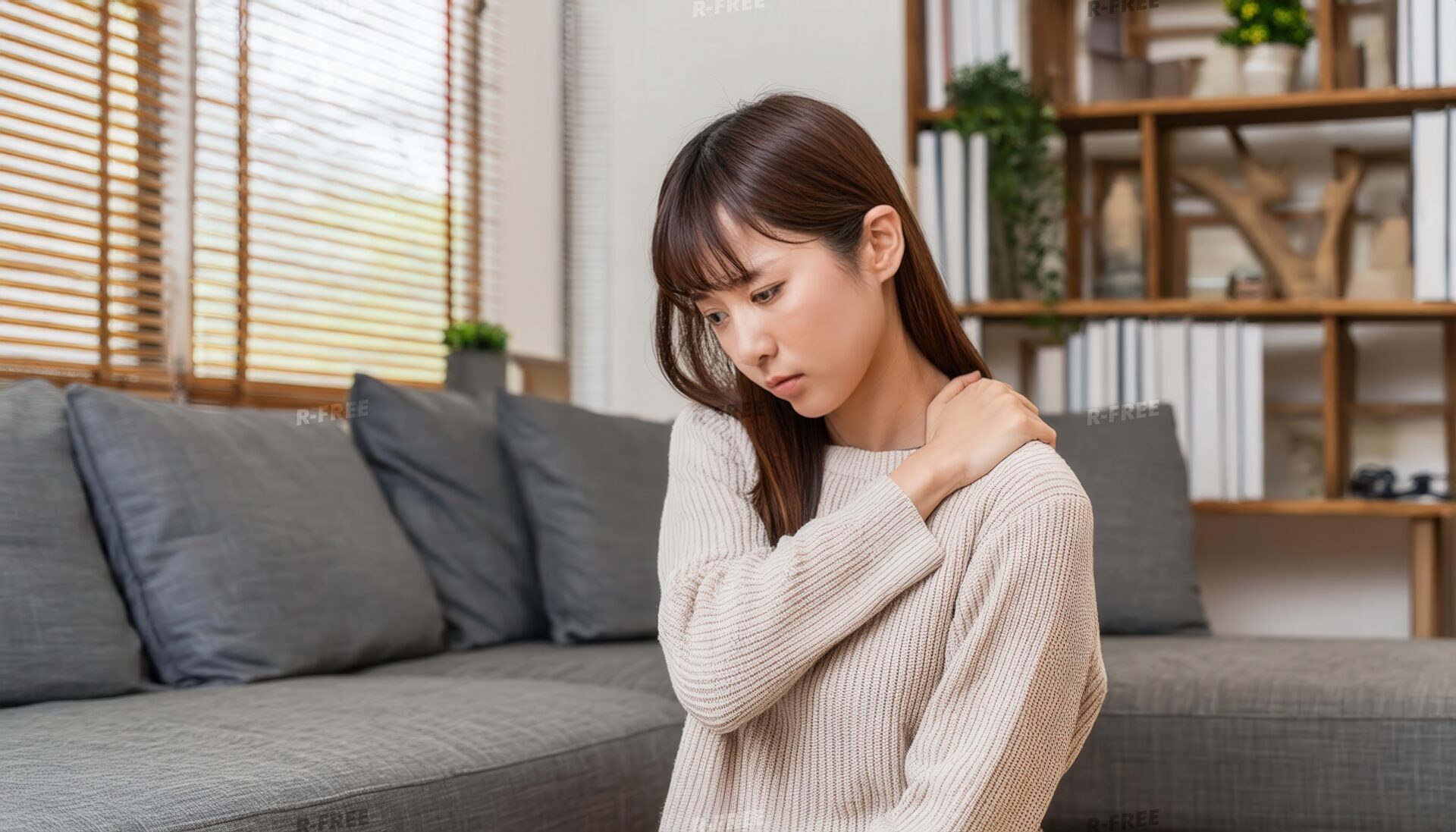 肩こりで左肩に手を添えて座るベージュニットの女性