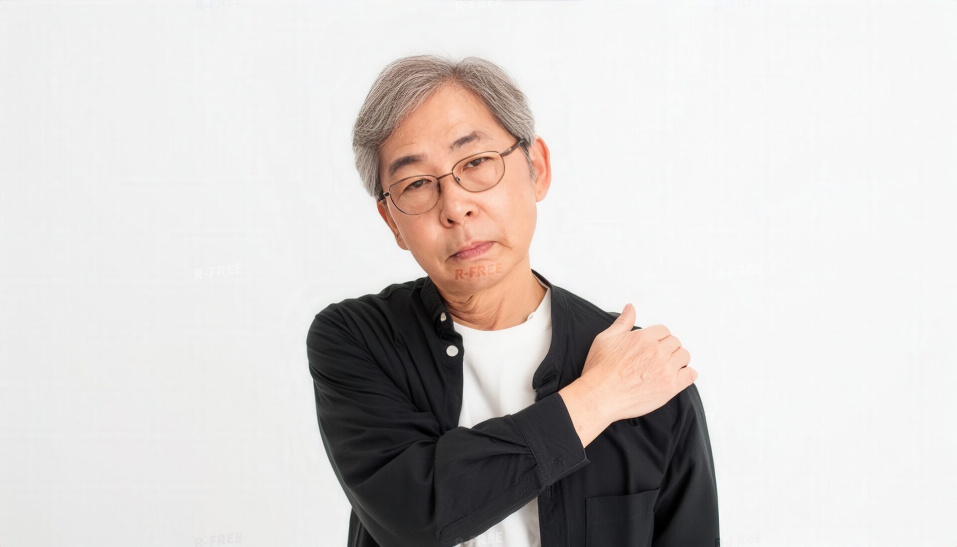 肩こりで左肩を押さえカメラを見る黒シャツのシニア男性