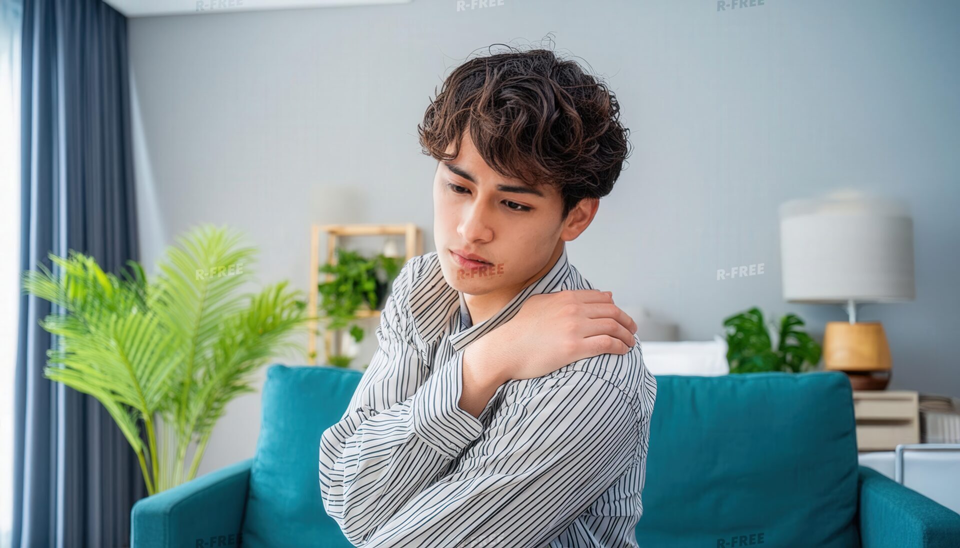 肩こりで左肩を押さえる青いソファの20代男性