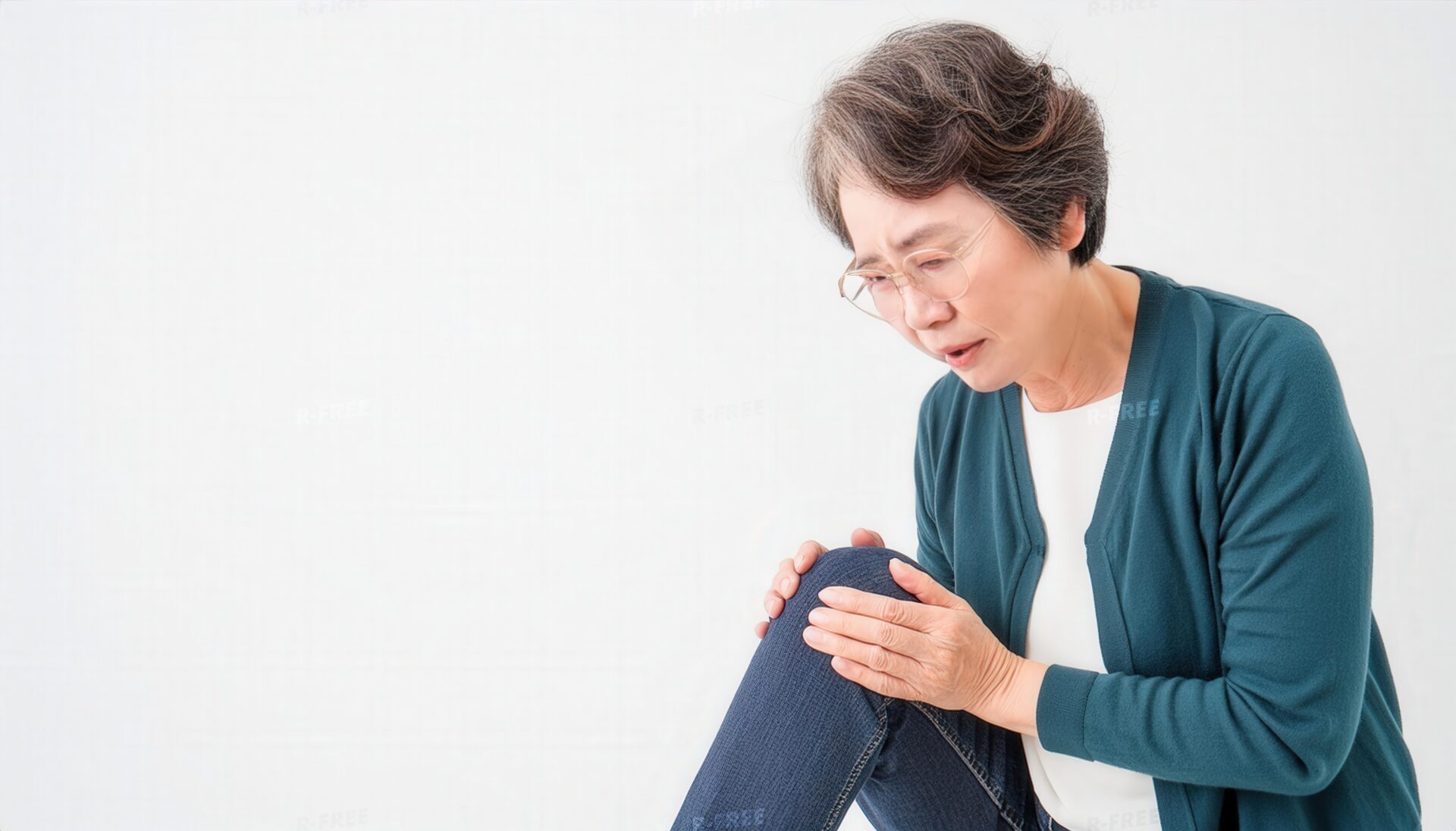 眼鏡と深緑カーディガン姿で前かがみに左膝を押さえる高齢女性