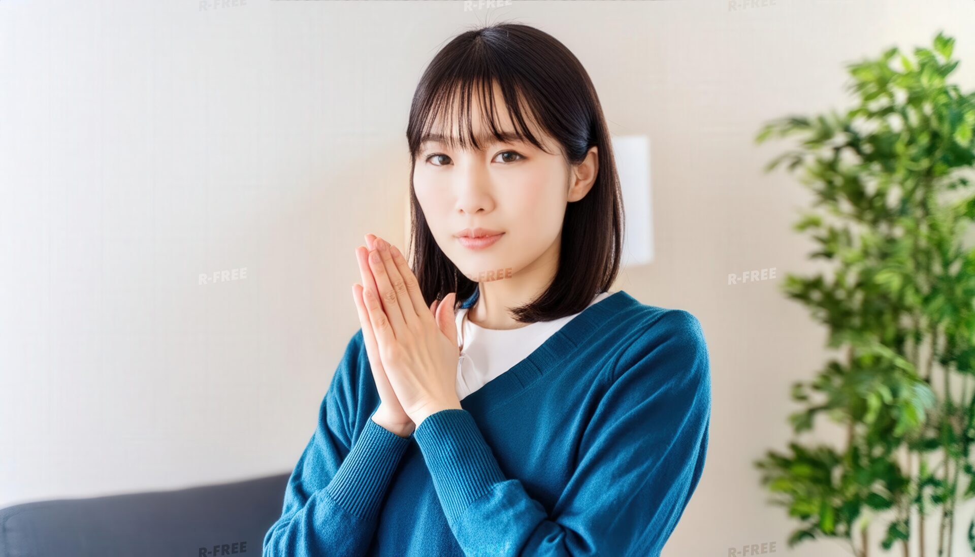 室内で胸元に手を合わせ正面を見る若い女性（ブルーセーター）