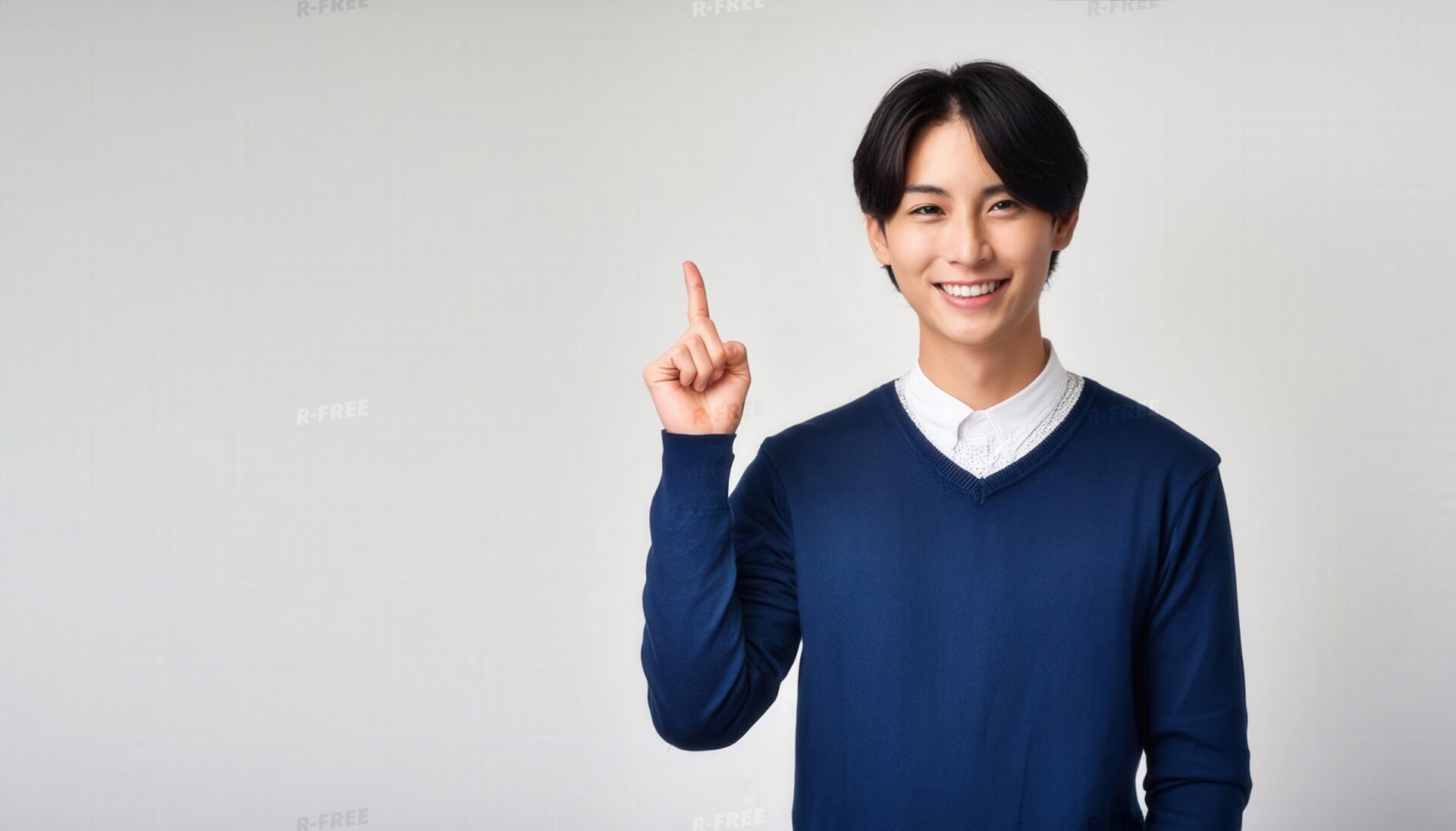 白背景で紺色のセーターを着て人差し指を立てる笑顔の若い男性