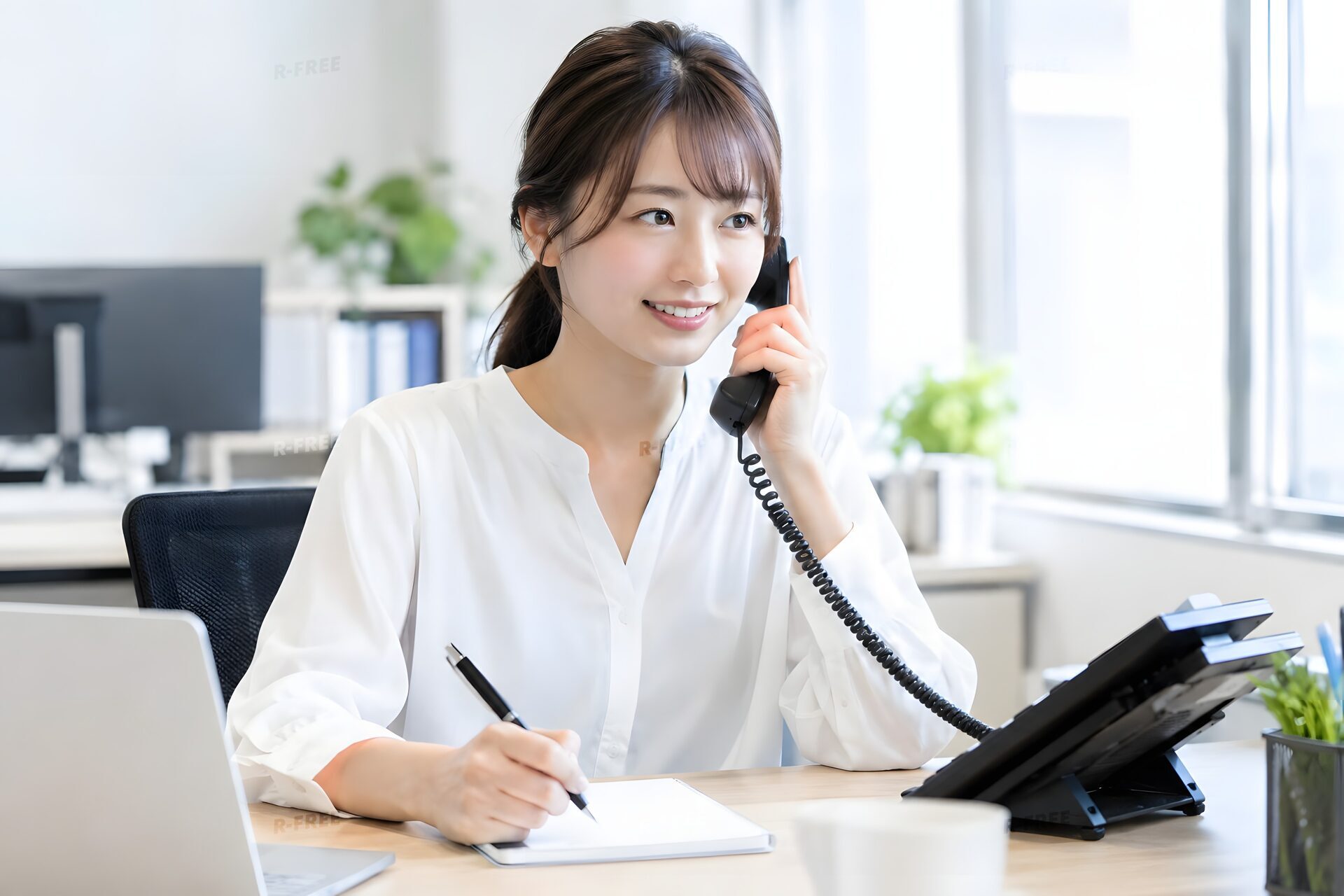オフィスの固定電話に笑顔で応対しメモを取る20代女性