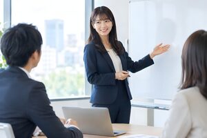 会議室でホワイトボードの前に立ち同僚に説明する若い女性会社員