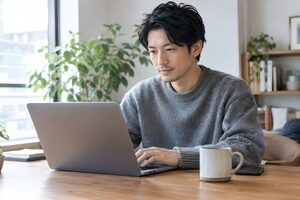 自宅の木製デスクでノートパソコンに向かう30代男性